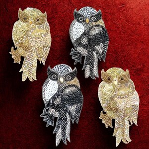 Op de afbeelding: Vier uilvormige broches met ingewikkelde kralen- en pailletten details. De broches hebben een verscheidenheid aan kleuren, waaronder goud, zilver, zwart en wit, tegen een dieprode achtergrond. Elke uil heeft een uniek ontwerp.