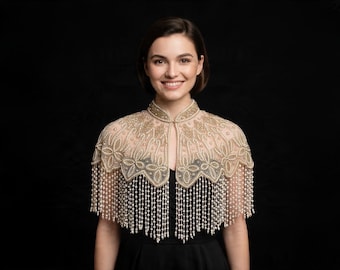 May include: A sheer, beige capelet featuring intricate gold embroidery and floral patterns, adorned with long, dangling strands of white pearls that create a fringe effect around the shoulders and chest, fastened at the high neckline.