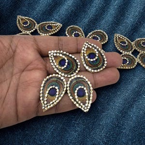 May include: A collection of teardrop-shaped patches featuring a peacock feather pattern in shades of blue, green, and gold, each bordered by a row of small, clear, reflective rhinestones. These decorative embellishments are arranged in groups.