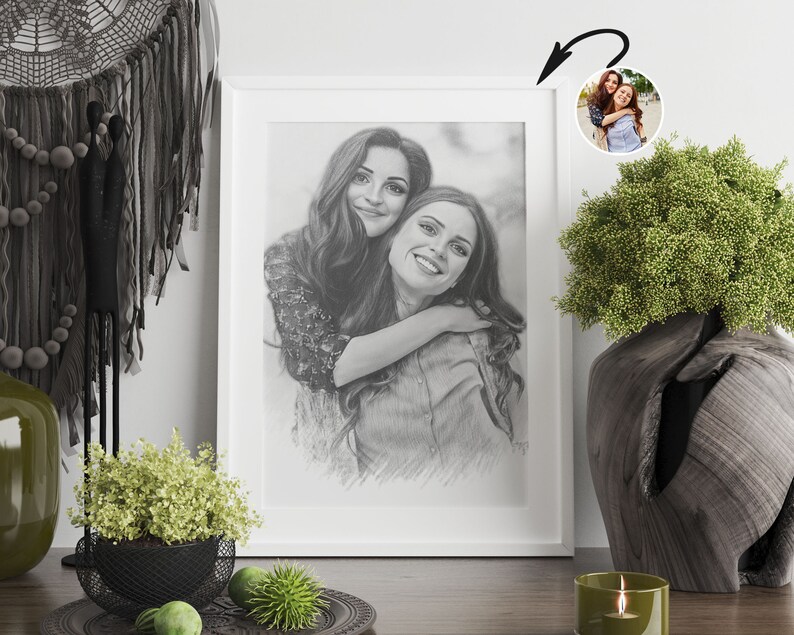 May include: A black and white pencil drawing of two women hugging. The drawing is framed in a white frame and is hanging on a white wall.