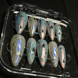 Puede incluir: Un conjunto de diez uñas postizas iridiscentes en forma de almendra. Cada uña presenta un diseño en espiral en tonos de azul, gris y blanco, acentuado con pequeños tachuelas plateadas. Las uñas se muestran en una caja de plástico transparente.
