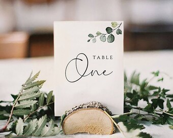 May include: A white card with a green watercolor eucalyptus branch design and the text "TABLE One" in black calligraphy. The card is sitting on a wooden slice.