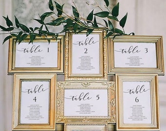 May include: Nine framed table number cards with elegant calligraphy, arranged on a wooden easel and adorned with greenery. Each card displays a table number and guest names, perfect for weddings or events.