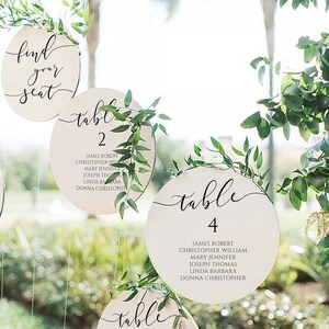 Peut inclure: Un ensemble de six pancartes rondes en bois numérotées de table avec des accents de verdure. Chaque pancarte présente une police d'écriture calligraphique noire pour le mot "table" et un numéro noir. Les pancartes sont pour un mariage ou un autre événement.