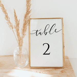 May include: A gold-framed table number sign with the word "table" and the number "2" in black calligraphy font. The sign is sitting on a wooden surface with a vase of dried pampas grass.