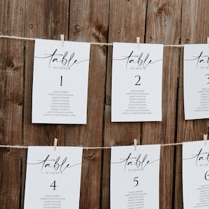 May include: Six white table number cards with black calligraphy. The cards are strung together with twine and clothespins. The table numbers are 1, 2, 3, 4, 5, and 6. Each card lists a group of names.