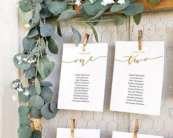 May include: Four white cards with gold calligraphy and text listing table numbers and guest names. The cards are attached to a wire mesh background with wooden clothespins. Greenery with white flowers is in the background.