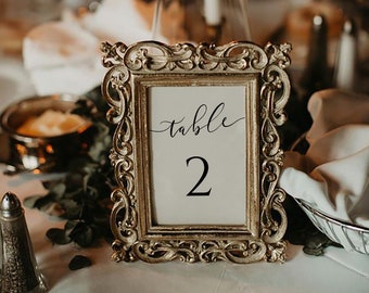 May include: Gold-colored ornate picture frame with a white card inside. The card has the text "table 2" written in black calligraphy.