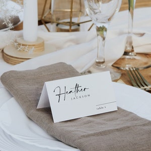 May include: A white place card with the name "Heather Jackson" and the text "table 1" written in black calligraphy. The place card is on a brown linen napkin, which is on top of a white plate.