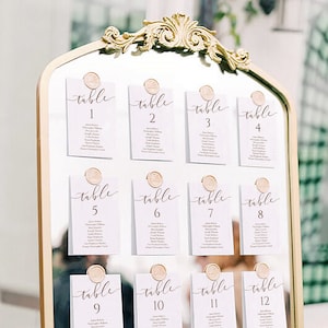 May include: A gold-framed mirror with white cards attached to it. The cards are labeled with the numbers 1 through 16 and the word "table" written in cursive. Each card has a list of names on it.