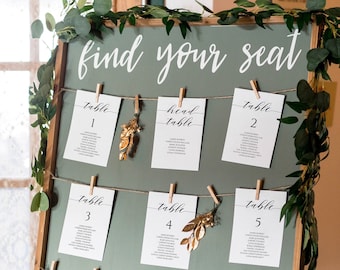 May include: A wooden easel with a green chalkboard displaying a wedding seating chart. The chalkboard has the text "find your seat" written in white cursive. The seating chart is organized by table number, with each table card listing the names of the guests assigned to that table. The cards are held in place by clothespins and decorated with gold leaf.