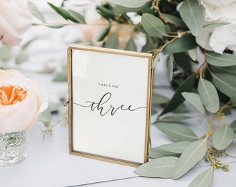 May include: A gold-framed table number sign with the word "three" written in cursive script. The sign is sitting on a light blue tablecloth with a bouquet of white and pink flowers and greenery behind it.