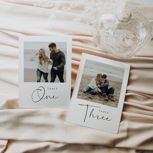 May include: Two white cards with black text and photos of a couple on a beach. The text on the cards reads "Table One" and "Table Three".