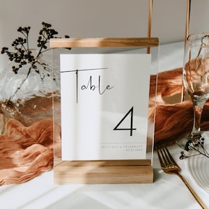 May include: A table number sign with a wooden base and clear acrylic panel. The sign displays the word "Table" in elegant script, the number "4", and the text "LET'S EAT & CELEBRATE 07.22.2025". The sign is set on a table with a gold fork.