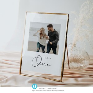 May include: A gold-framed table number sign with a white background. The sign features a photo of a couple walking on a beach and the text "TABLE One" in a cursive font.