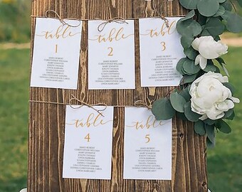 May include: A rustic wooden wedding seating chart with greenery and white flowers. The chart has five cards with the words "table" and numbers 1 through 5. Each card lists guest names.