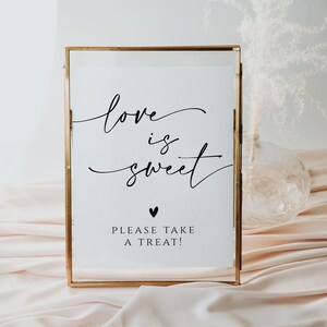 May include: A gold framed sign with the text "love is sweet" in black calligraphy. Below the text is a small black heart and the text "Please take a treat!"