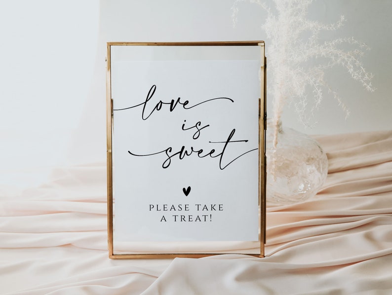May include: A gold framed sign with the text "love is sweet" in black calligraphy. Below the text is a small black heart and the text "Please take a treat!"