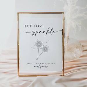 May include: A white sign in a gold frame with the text "Let love sparkle" and a graphic of two sparklers with hearts at the top. The text below the graphic reads "Light the way for the newlyweds".