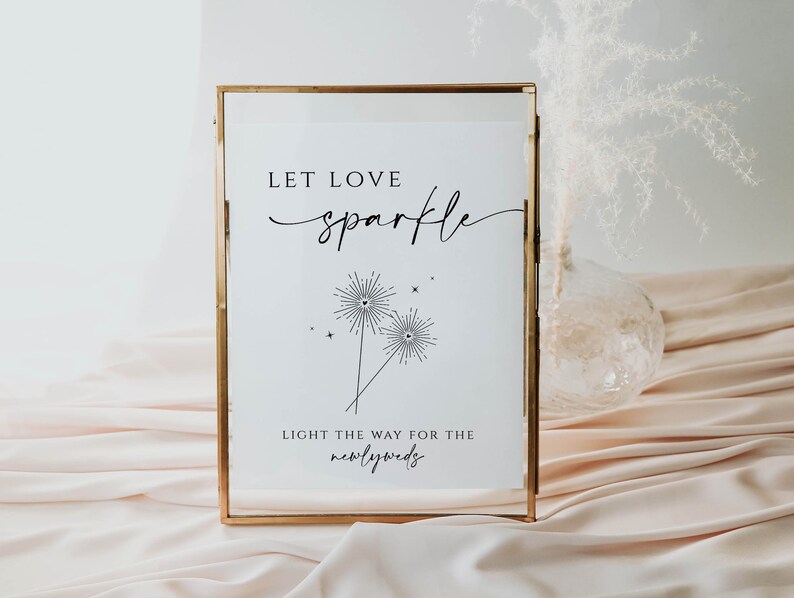 May include: A white sign in a gold frame with the text "Let love sparkle" and a graphic of two sparklers with hearts at the top. The text below the graphic reads "Light the way for the newlyweds".