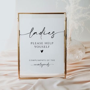May include: A gold-framed sign with the text "ladies please help yourself" and a small heart symbol. Below the text is "compliments of the newlyweds".
