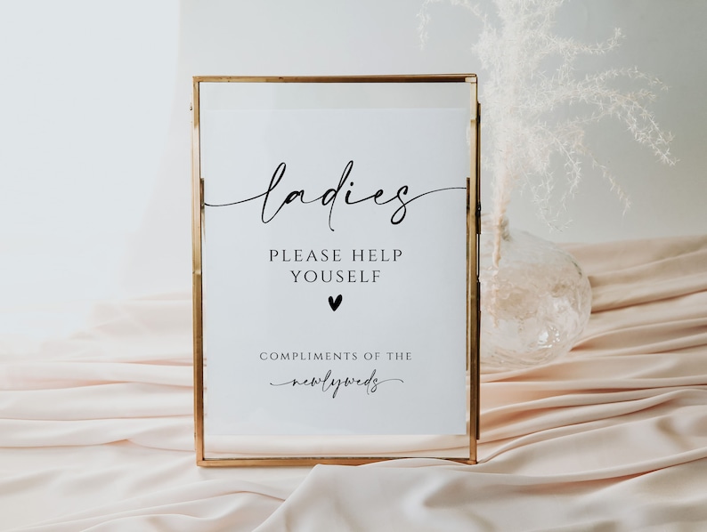 May include: A gold-framed sign with the text "ladies please help yourself" and a small heart symbol. Below the text is "compliments of the newlyweds".