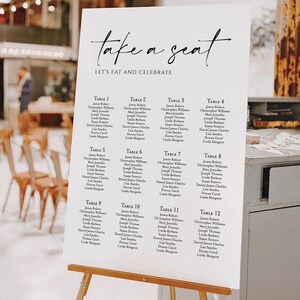 K&ouml;nnte beinhalten: Ein wei&szlig;es Hochzeits-Sitzplan auf einer Holzstaffelei. Die Tafel zeigt eine elegante Kalligraphie mit der Aufschrift "take a seat" und "let's eat and celebrate". Darunter sind 12 Tische mit den Namen der G&auml;ste aufgef&uuml;hrt. Der Hintergrund ist ein verschwommenes Interieur.