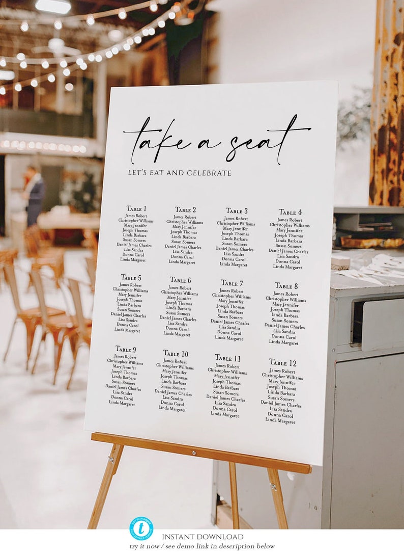 K&ouml;nnte beinhalten: Ein wei&szlig;es Hochzeits-Sitzplan auf einer Holzstaffelei. Die Tafel zeigt eine elegante Kalligraphie mit der Aufschrift "take a seat" und "let's eat and celebrate". Darunter sind 12 Tische mit den Namen der G&auml;ste aufgef&uuml;hrt. Der Hintergrund ist ein verschwommenes Interieur.