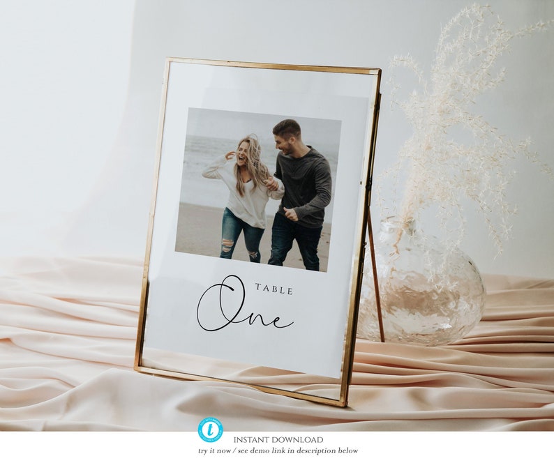 May include: A gold-framed table number sign with a white background. The sign features a photo of a couple walking on a beach and the text "TABLE One" in a cursive font.