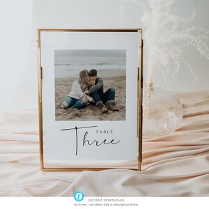 May include: A gold-framed table number sign with a photo of a couple on a beach. The sign reads "Table Three" in a modern calligraphy font.