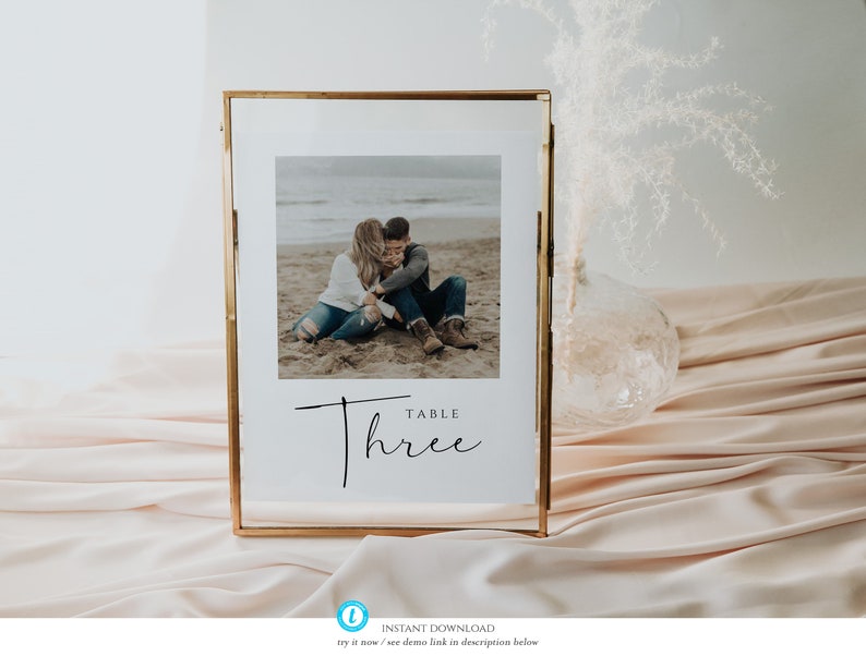 May include: A gold-framed table number sign with a photo of a couple on a beach. The sign reads "Table Three" in a modern calligraphy font.