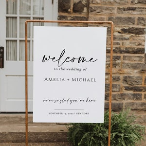 K&ouml;nnte beinhalten: Ein wei&szlig;es Hochzeits-Willkommensschild mit schwarzem Kalligraphie-Text. Auf dem Schild steht "welcome to the wedding of AMELIA + MICHAEL" und "we're so glad you're here". Das Schild wird von einem kupferfarbenen Metallrahmen gehalten. Datum und Ort sind ebenfalls angegeben.