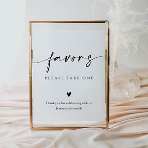 May include: A gold framed sign with the word "favors" written in cursive script. The sign also says "Please take one" and "Thank you for celebrating with us! It means the world!"