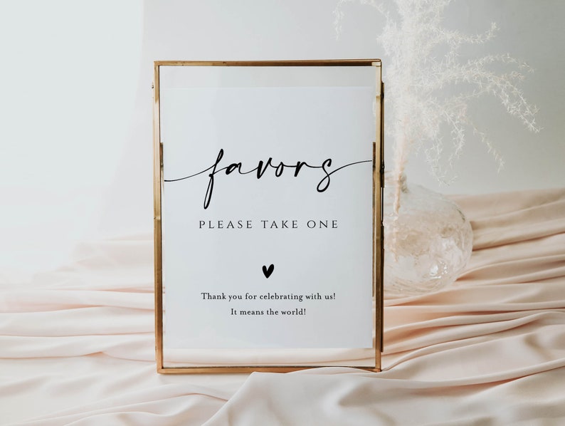 May include: A gold framed sign with the word "favors" written in cursive script. The sign also says "Please take one" and "Thank you for celebrating with us! It means the world!"