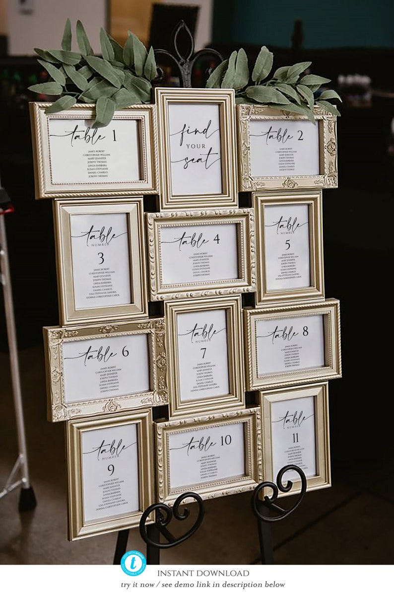 May include: A gold picture frame seating chart with 11 frames, each with a white card listing a table number and guest names. The frames are arranged in a grid pattern and are decorated with greenery.