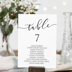 May include: A white table number card with the word "table" in elegant black script and the number 7. Below, guest names are listed. The card is held in a small wooden holder, with greenery and a vase in the background.