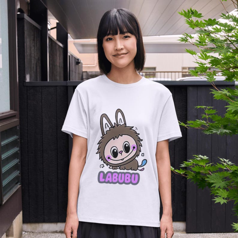 May include: White t-shirt featuring a cartoon character with bunny ears and a furry body. The character has a smiling face with large eyes and a blue tail. The word "LABUBU" is printed in purple below the character.