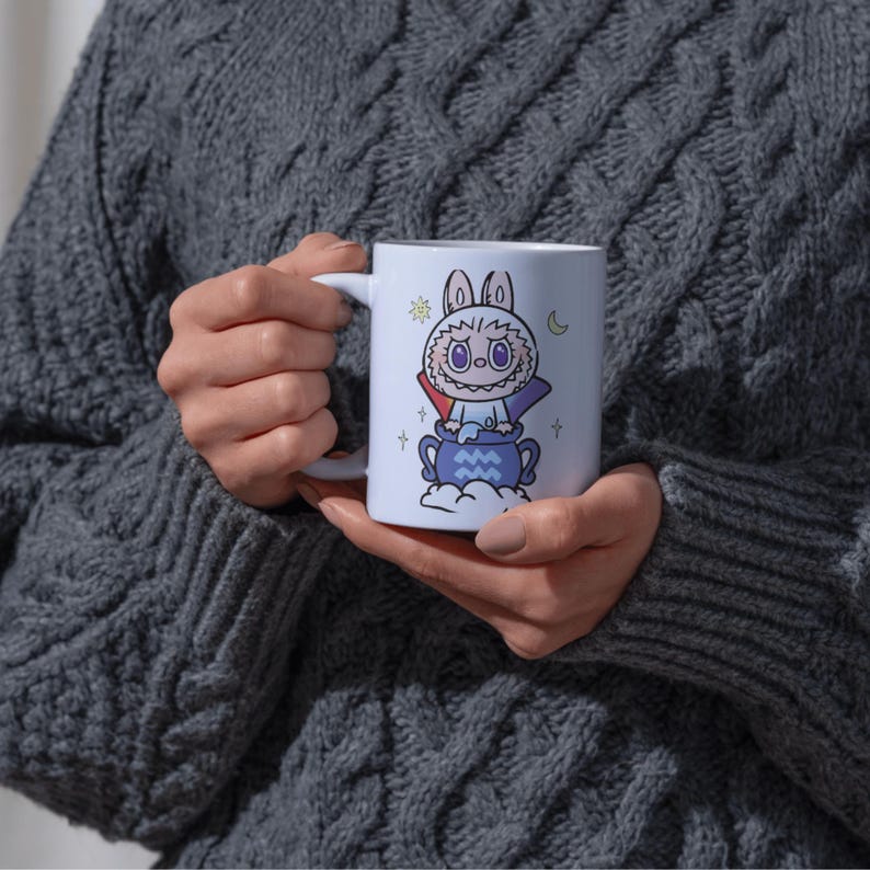 May include: White ceramic mug featuring a cartoon illustration of a bunny-like creature with a purple cape, a blue cauldron, and celestial accents. The mug has a white handle and is held by a person wearing a gray sweater.