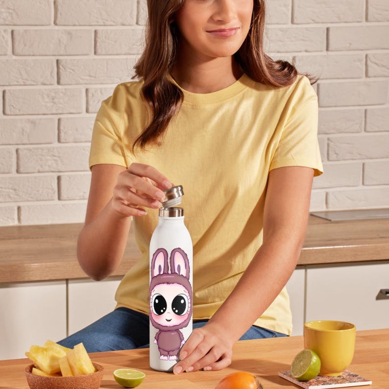 May include: A white water bottle featuring a cartoon bunny design in shades of purple and pink. The bottle has a silver cap and is being opened. The background includes a wooden surface, a yellow cup, and a bowl of pineapple.