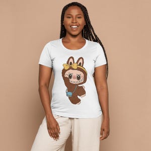 May include: White t-shirt featuring a cartoon character in a brown furry costume with a yellow bow and a small blue purse. The character has large eyes and a wide smile. The shirt is a crew neck style.
