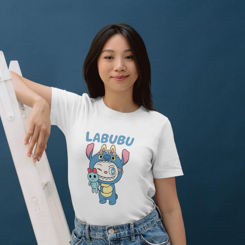 May include: White t-shirt featuring a cartoon character in a blue costume with the word "LABUBU" in light blue above the character. The character is holding a small blue doll with a red bow. The background is a solid dark blue.