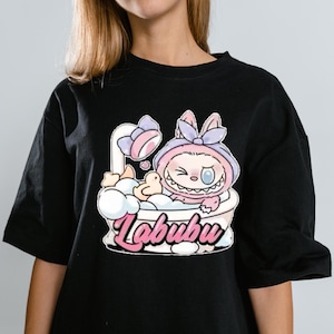 May include: Black t-shirt featuring a cartoon character in a bathtub with a shower head. The character is wearing a pink hooded outfit with a bow. The word "Labubu" is written in pink script below the bathtub.