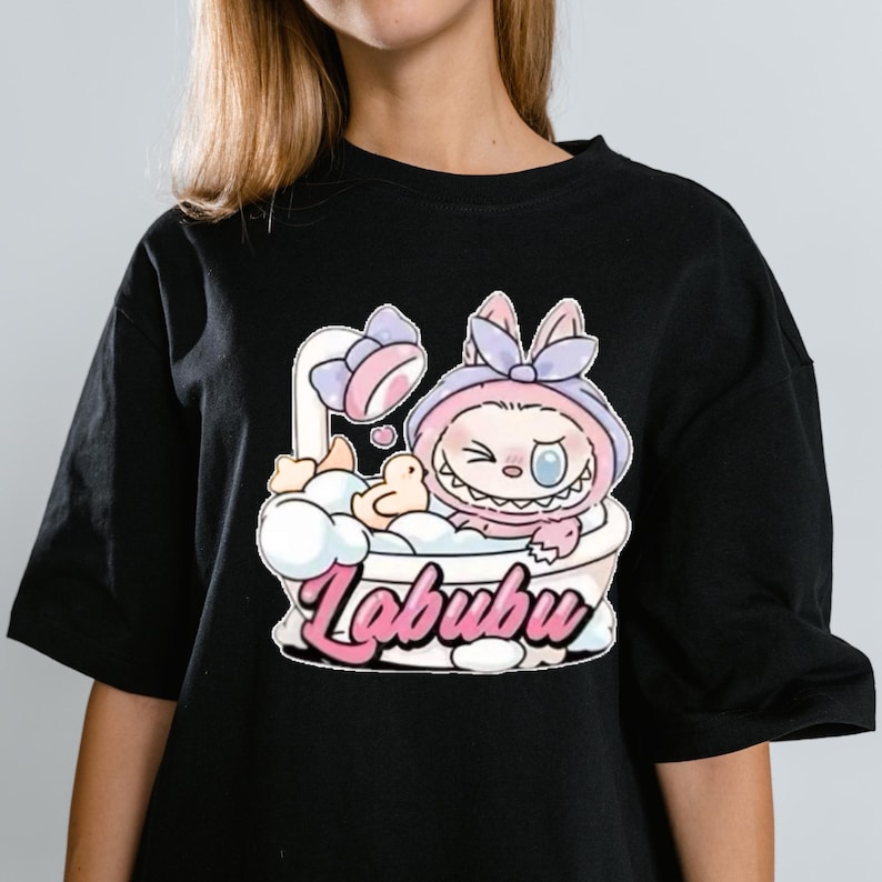 May include: Black t-shirt featuring a cartoon character in a bathtub with a shower head. The character is wearing a pink hooded outfit with a bow. The word "Labubu" is written in pink script below the bathtub.