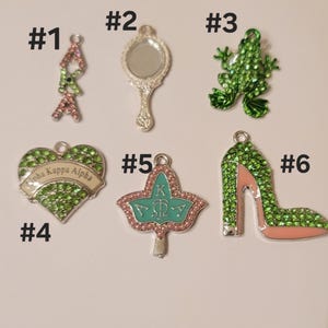 May include: Six different charms with green and pink rhinestones. The charms include a heart with the text "Alpha Kappa Alpha", a frog, a mirror, a leaf, a high heel shoe, and a letter "A".