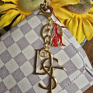 May include: A gold-toned metal keychain featuring a large stylized letter charm, a smaller rhinestone-encrusted circular charm, and a red enameled figure charm, all attached to a gold-toned lobster clasp and ring, resting on a white and brown checkered bag.