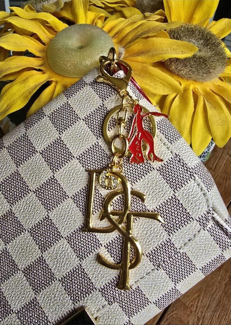May include: A gold-toned metal keychain featuring a large stylized letter charm, a smaller rhinestone-encrusted circular charm, and a red enameled figure charm, all attached to a gold-toned lobster clasp and ring, resting on a white and brown checkered bag.