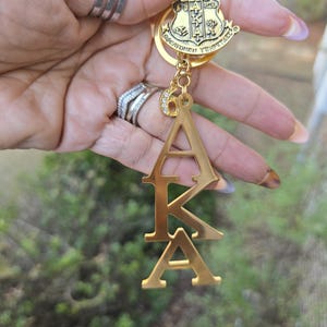 May include: A gold-toned metal keychain featuring a shield-shaped crest with the Greek letters AKA, a small charm with the number 6 encrusted with clear rhinestones, and a vertical pendant displaying the Greek letters A, K, and A in a stacked design.