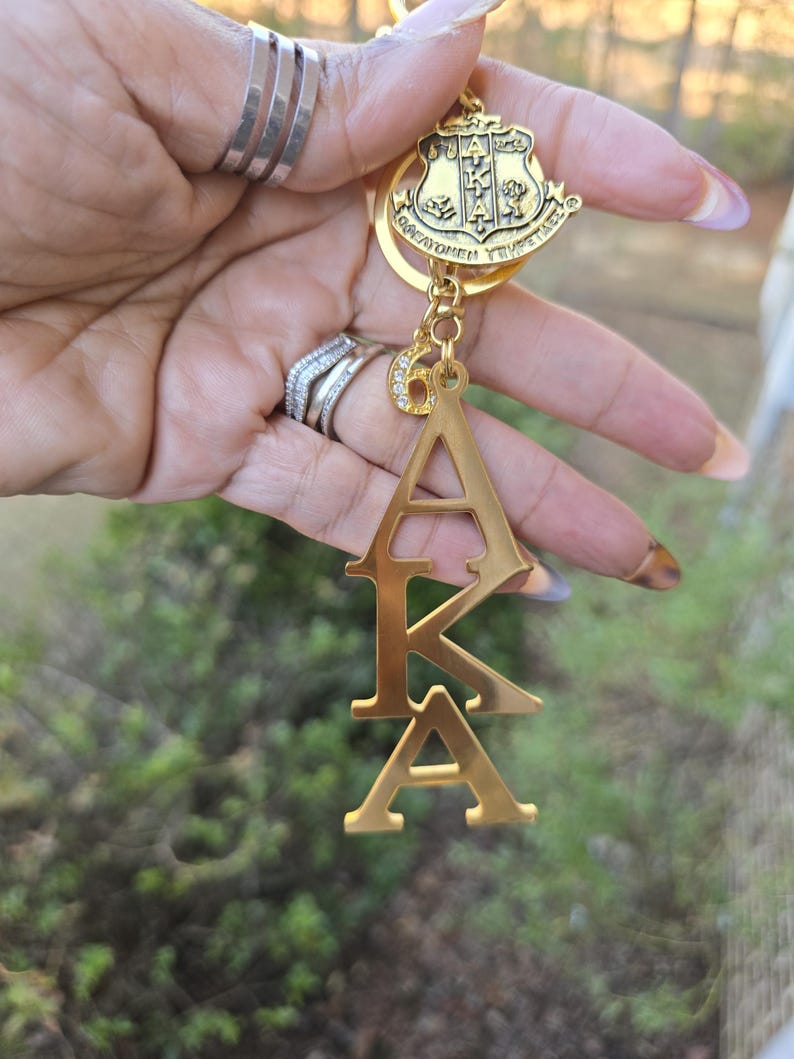 May include: A gold-toned metal keychain featuring a shield-shaped crest with the Greek letters AKA, a small charm with the number 6 encrusted with clear rhinestones, and a vertical pendant displaying the Greek letters A, K, and A in a stacked design.