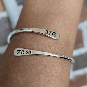 May include: A silver-colored cuff bracelet with the Greek letters &Delta;&Sigma;&Theta; engraved on one end and "SPR '26" on the other. The bracelet is curved and open, designed to be worn on the wrist. The background is a blurred view of a brick wall.