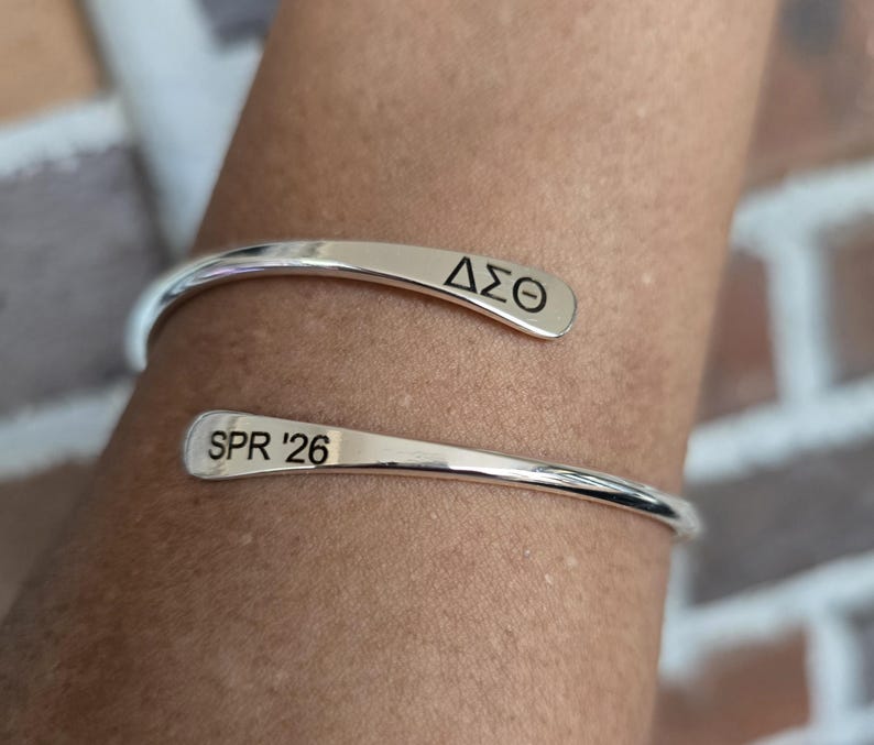 May include: A silver-colored cuff bracelet with the Greek letters &Delta;&Sigma;&Theta; engraved on one end and "SPR '26" on the other. The bracelet is curved and open, designed to be worn on the wrist. The background is a blurred view of a brick wall.